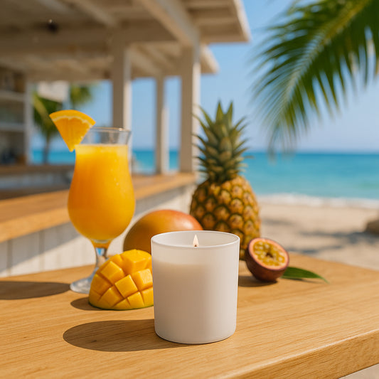 Eine sommerliche Szene am Strand mit Blick auf eine Strandbar und das glitzernde Meer. Auf einem Holztisch steht eine brennende Duftkerze im mattweiß beschichteten Glas, flankiert von frischer Mango, Ananas und Passionsfrucht. Daneben ein Cocktail mit Orangenscheibe als Dekoration. Palmenblätter werfen weiche Schatten auf den Tisch.